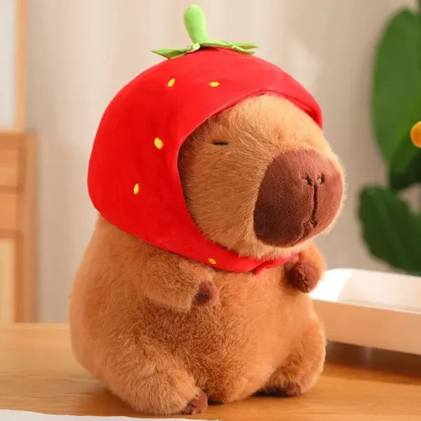 Peluche capybara brun assis yeux fermés, coiffé d'un chapeau fraise rouge kawaii sur bois clair.